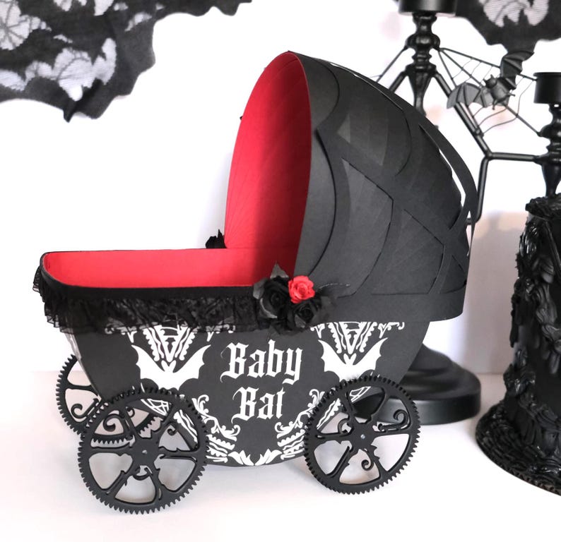 May include: A decorative black and red baby carriage featuring the words "Baby Bat" in white gothic script. The carriage has gear-shaped wheels and is decorated with black lace and a red rose. The carriage is approximately 30 cm long.