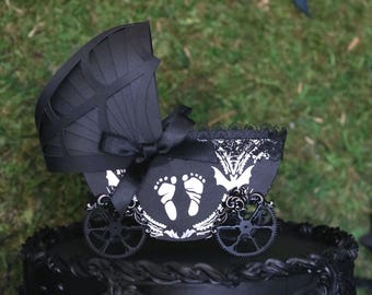 May include: A black baby carriage cake topper with a decorative design. The carriage features a black bow, white footprints, and a decorative wheel. The carriage is on top of a black cake with an ornate design. The background is green grass.