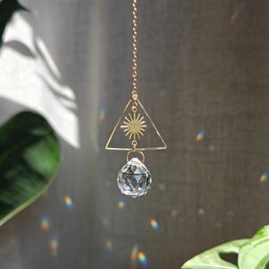 May include: A gold-toned metal suncatcher featuring a delicate chain, a triangular frame with a central sunburst emblem, and a clear, multifaceted crystal sphere hanging from the bottom, designed to refract light into small rainbow patterns.