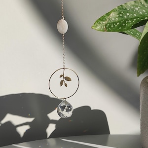 Op de afbeelding: Een decoratief hangornament met een heldere kristallen bol, een goudkleurige metalen ring met bladeren en een witte steen. Het ornament hangt aan een gouden ketting. Een kleine, getextureerde, beige vaas bevat een groene plant met witte stippen.