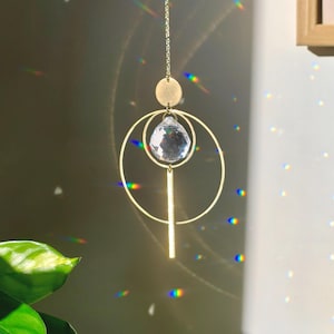Può includere: Un acchiappasole in metallo color oro con una cornice circolare, una barra verticale e un piccolo disco solido, sospeso da una catena sottile. Un prisma di cristallo trasparente sfaccettato è centrato all'interno degli anelli.