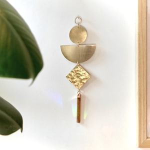Op de afbeelding: Een verticale wandhanger bestaande uit een kleine messing cirkel, een messing halve cirkel en een gehamerde messing ruitvorm verbonden door metalen ringen, eindigend in een rechthoekig bruin stenen prisma dat licht vangt voor een regenboogeffect.
