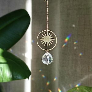 Può includere: Un ciondolo in metallo dorato a forma di sole sospeso a una catenina sottile, caratterizzato da una cornice circolare con un motivo radiante centrale. Un cristallo trasparente sfaccettato a goccia pende dalla parte inferiore dell'anello.