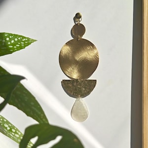 Peut inclure: Une décoration murale suspendue composée d'un petit cercle en laiton, d'un grand cercle en laiton brossé, d'un demi-cercle en laiton martelé et d'un pendentif en forme de goutte blanc translucide, reliés verticalement par des anneaux.