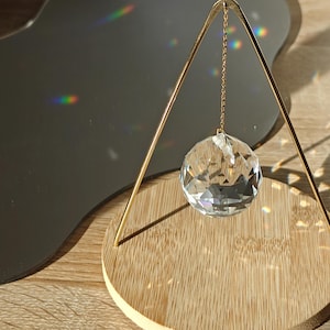 May include: A clear crystal ball suspended by a gold-coloured metal frame and chain, set on a round bamboo base. A dark grey, wavy-edged mirror and wooden surface form the background. Rainbow light reflections are visible.