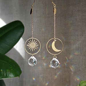 May include: Two gold-toned hanging ornaments suspended from a thin metal rod. One features a sun emblem inside a circular frame, the other a crescent moon. Both hang above a clear, faceted, teardrop-shaped crystal prism that reflects light.