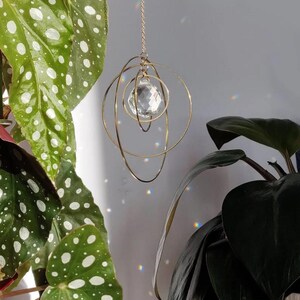 May include: A hanging suncatcher featuring a central faceted clear crystal sphere suspended within three interlocking gold-toned metal rings. The ornament hangs from a thin gold-toned chain, casting small rainbow light refractions on the wall.