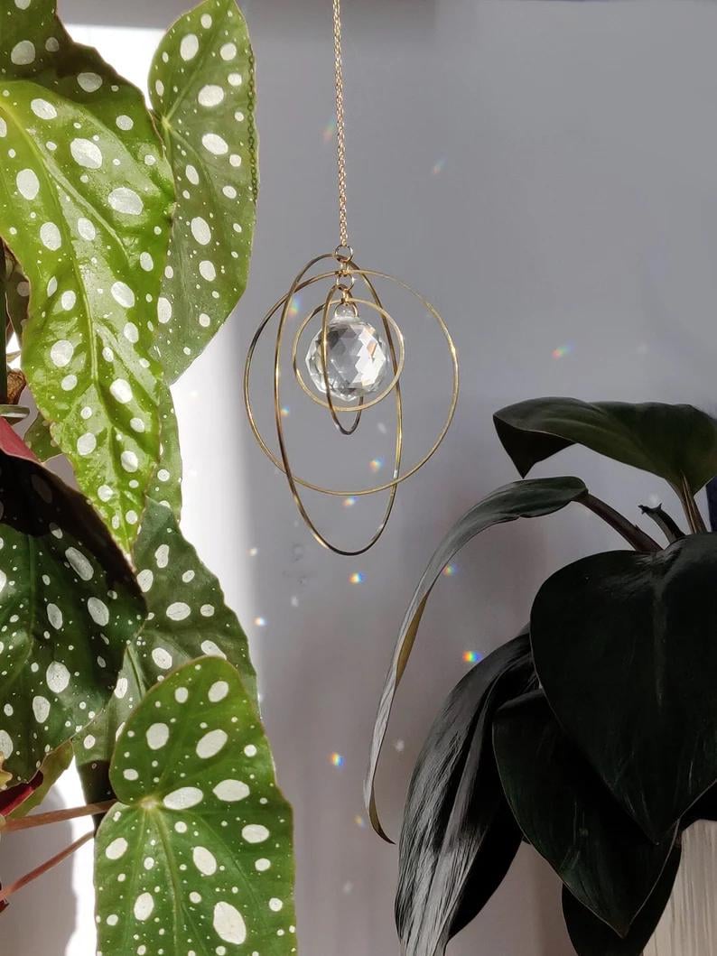 May include: A hanging suncatcher featuring a central faceted clear crystal sphere suspended within three interlocking gold-toned metal rings. The ornament hangs from a thin gold-toned chain, casting small rainbow light refractions on the wall.