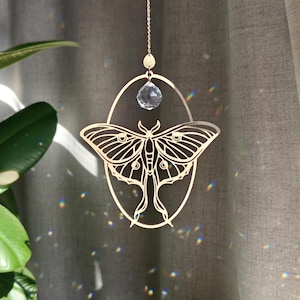 Peut inclure: Un attrape-soleil en métal doré présentant un motif de papillon de nuit découpé au centre d'un cadre ovale. Une perle de cristal transparente à facettes est suspendue au sommet de l'ovale par une fine chaîne dorée vers le haut.