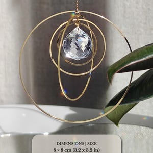 May include: A hanging ornament featuring three concentric gold-toned metal rings and a faceted clear crystal prism. The total dimensions are 8 x 8 cm, with a 2.5 cm crystal. The design is suspended by a thin gold chain, creating light reflections.