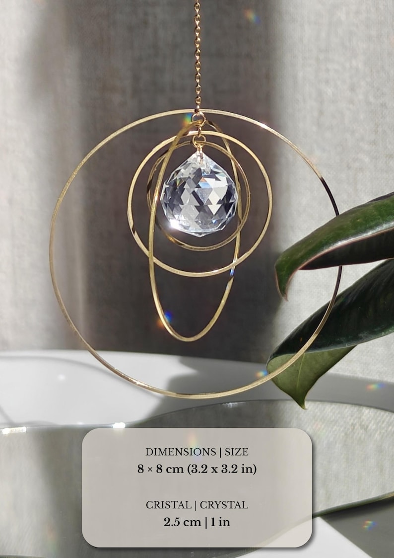 May include: A hanging ornament featuring three concentric gold-toned metal rings and a faceted clear crystal prism. The total dimensions are 8 x 8 cm, with a 2.5 cm crystal. The design is suspended by a thin gold chain, creating light reflections.