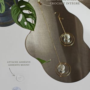 May include: Two gold-toned metal chains with circular pendants featuring clear crystal spheres. Includes a brass hook, adhesive mount, and suction cup for hanging. Text reads: BRASS HOOK INCLUDED, ATTACHE ADHESIVE ADHESIVE MOUNT, VENTOUSE SUCTION CUP, FACILE &Agrave; SUSPENDRE EASY TO HANG.