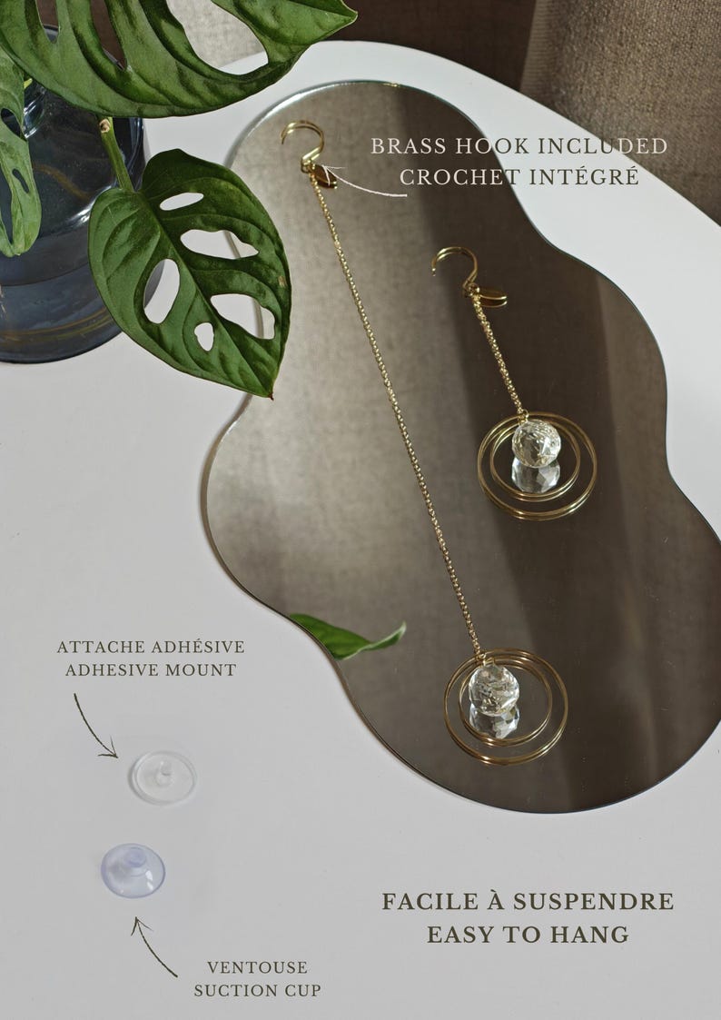 May include: Two gold-toned metal chains with circular pendants featuring clear crystal spheres. Includes a brass hook, adhesive mount, and suction cup for hanging. Text reads: BRASS HOOK INCLUDED, ATTACHE ADHESIVE ADHESIVE MOUNT, VENTOUSE SUCTION CUP, FACILE &Agrave; SUSPENDRE EASY TO HANG.