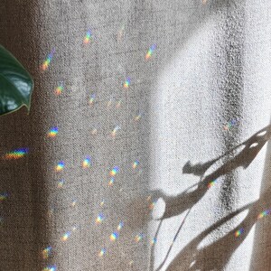 May include: A textured beige fabric curtain displays scattered prismatic light reflections in a spectrum of colours. A dark green leaf is visible on the left side, and a distinct shadow of a plant leaf is cast onto the fabric surface.