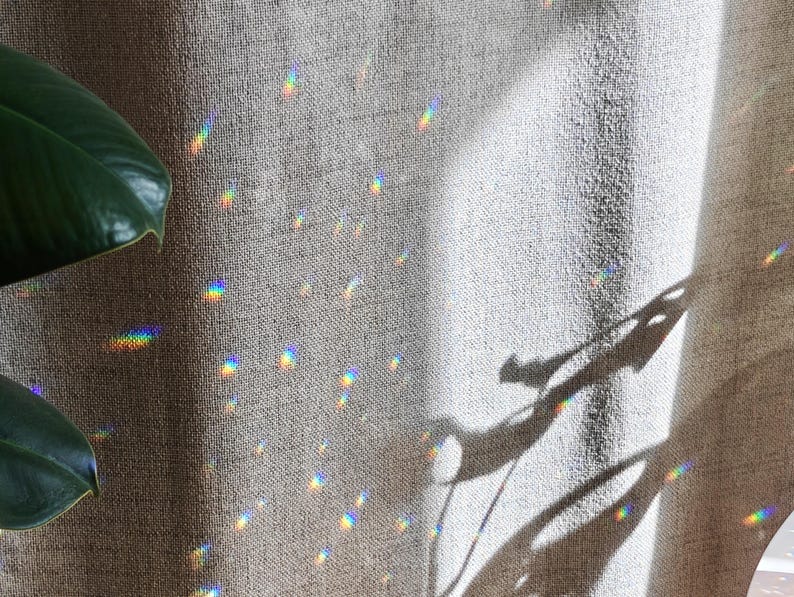 May include: A textured beige fabric curtain displays scattered prismatic light reflections in a spectrum of colours. A dark green leaf is visible on the left side, and a distinct shadow of a plant leaf is cast onto the fabric surface.