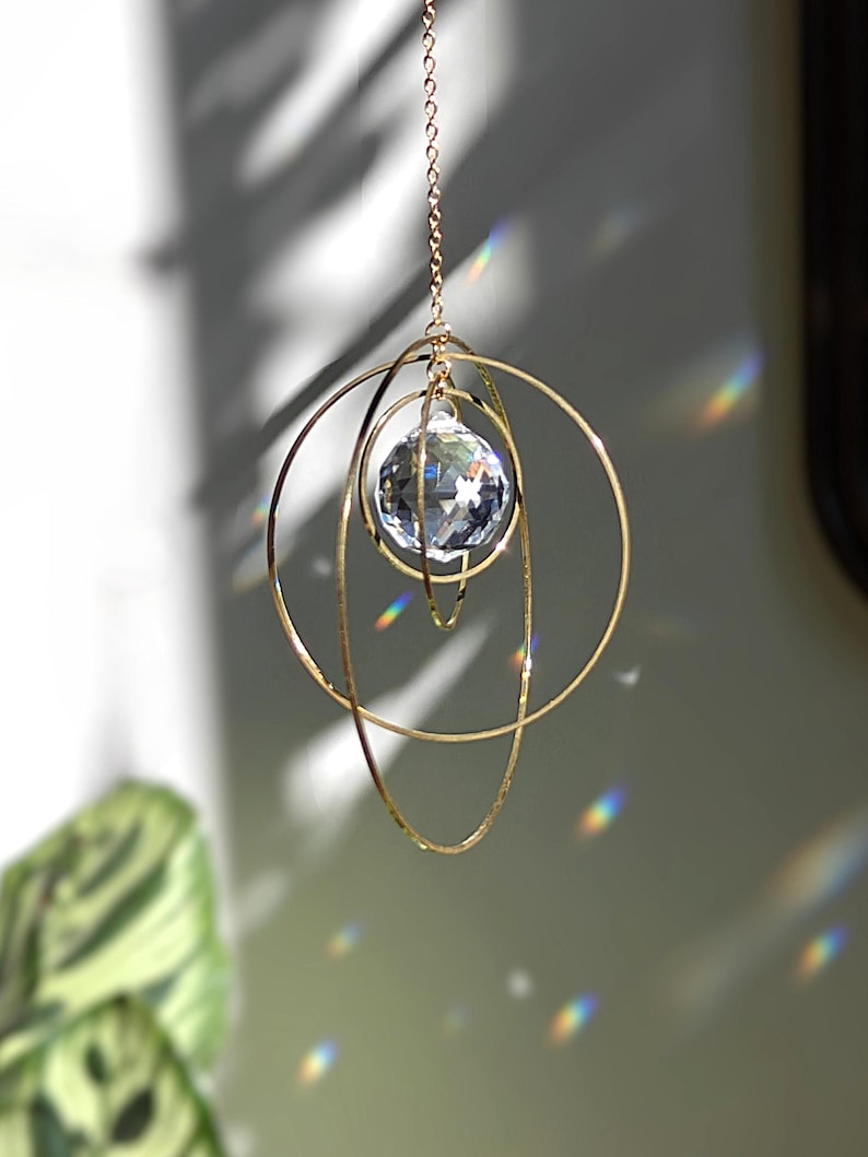 May include: A hanging suncatcher featuring a clear, multifaceted crystal sphere suspended within three interlocking gold-toned metal rings. The structure is attached to a thin gold chain, and light passing through the crystal creates rainbow refractions.