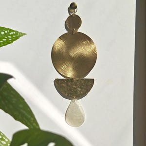 May include: A vertical wall hanging ornament composed of a small brass circle, a large brushed brass circle, a hammered brass semi-circle, and a translucent white teardrop-shaped pendant, all connected by small metal jump rings in a linear arrangement.