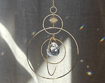 May include: A gold-toned metal suncatcher featuring a series of concentric and overlapping circular rings suspended from a delicate chain. A faceted clear crystal prism hangs in the center, catching light to create small rainbow refractions.