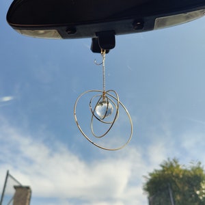 May include: A hanging car ornament featuring a clear, multifaceted crystal sphere suspended by a thin gold-toned chain, surrounded by three interlocking gold-toned metal rings of varying sizes that create a spherical, orbital-like structure.