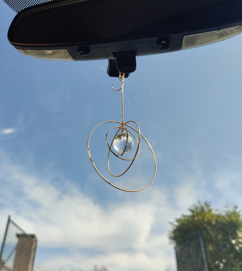 May include: A hanging car ornament featuring a clear, multifaceted crystal sphere suspended by a thin gold-toned chain, surrounded by three interlocking gold-toned metal rings of varying sizes that create a spherical, orbital-like structure.