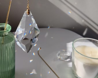 May include: A clear crystal prism hanging from a gold chain, casting rainbow light on a white surface. A green glass vase and a candle are also visible.