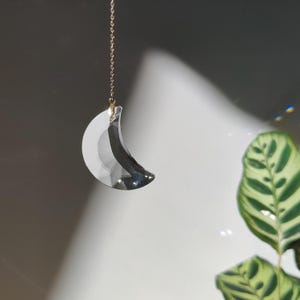 Pode incluir: Um pingente de lua de cristal transparente e facetado, pendurado por uma delicada corrente dourada. O apanhador de sol em forma de crescente reflete a luz, criando um efeito cintilante. O fundo é cinza suave com um toque de verde de uma planta próxima.