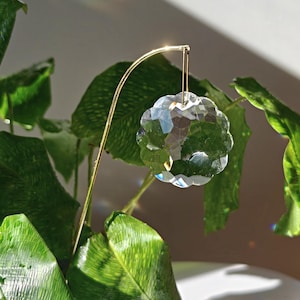 Könnte beinhalten: Ein klarer, facettierter Kristall-Sonnenfänger in Blumenform, der an einem dünnen, geschwungenen goldfarbenen Metalldrahtständer hängt. Der Kristall reflektiert das Licht und erzeugt kleine Regenbogenprismen auf den grünen Blättern.