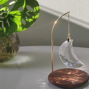 Peut inclure: Un pendentif en cristal transparent en forme de croissant de lune à facettes est suspendu à un fin support en métal doré. Le support est fixé sur une base circulaire en bois brun foncé. La lune en cristal reflète la lumière sur la base.