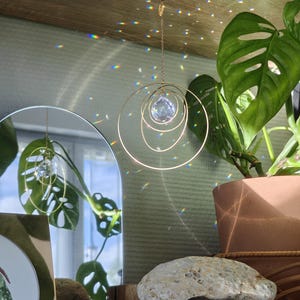 May include: A hanging suncatcher featuring a faceted clear crystal sphere suspended within three concentric gold-toned metal rings. The ornament is attached to a thin gold chain and a small hook, casting colourful light refractions on the surrounding area.
