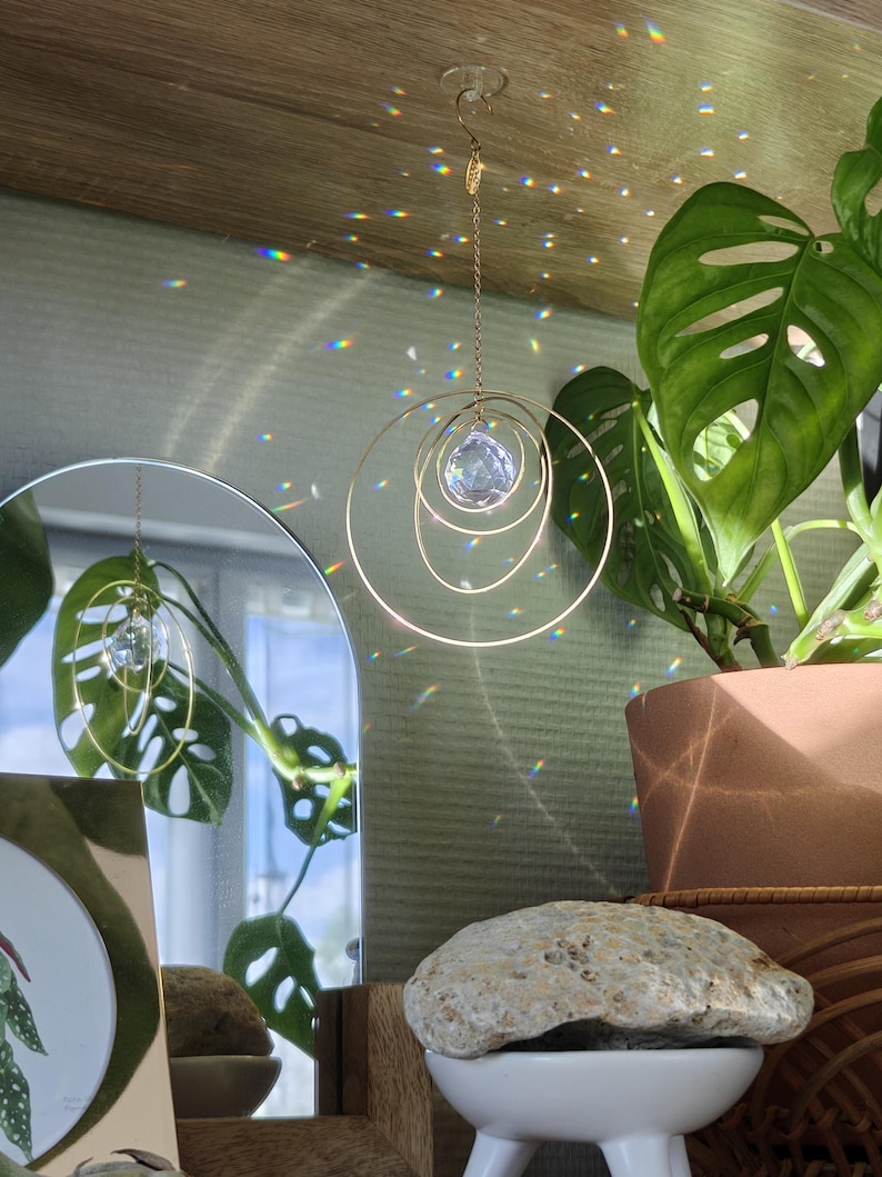 May include: A hanging suncatcher featuring a faceted clear crystal sphere suspended within three concentric gold-toned metal rings. The ornament is attached to a thin gold chain and a small hook, casting colourful light refractions on the surrounding area.