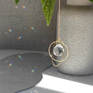 Könnte beinhalten: Ein goldfarbener Sonnenfänger aus Metall mit einer facettierten, klaren Kristallkugel, die in zwei ineinandergreifenden goldenen Ringen hängt. Das Ornament ist an einer feinen goldenen Kette befestigt und wirft kleine Regenbogenlichtreflexe.