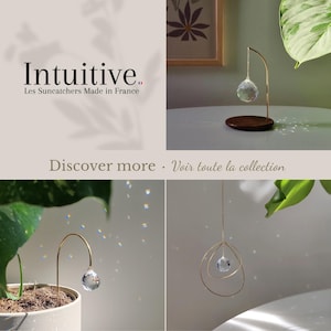 May include: A collection of suncatchers featuring clear faceted crystal prisms suspended from gold-toned metal stands and hoops. One design includes a dark wood circular base. Text reads: Intuitive. Les Suncatchers Made in France. Discover more.