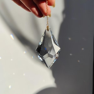 Op de afbeelding: Een helder, facetgeslepen kristallen prisma, diamantvormig, opgehangen aan een fijne gouden ketting. Het kristal reflecteert licht, waardoor een sprankelend effect ontstaat. Het prisma wordt tegen een witte achtergrond gehouden, met schaduwen en lichtreflecties.