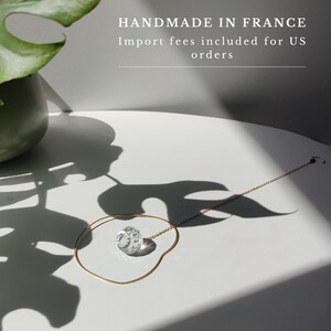 May include: A delicate gold-toned chain necklace featuring a single, clear, multifaceted spherical crystal pendant. The necklace is arranged in a circular shape on a white surface, accompanied by the text HANDMADE IN FRANCE.