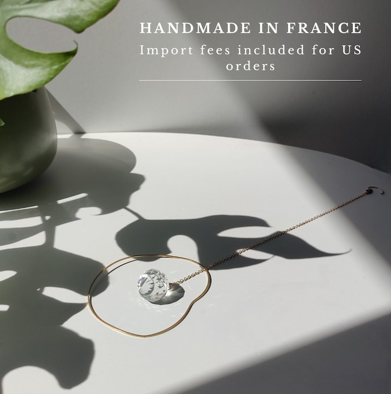 May include: A delicate gold-toned chain necklace featuring a single, clear, multifaceted spherical crystal pendant. The necklace is arranged in a circular shape on a white surface, accompanied by the text HANDMADE IN FRANCE.