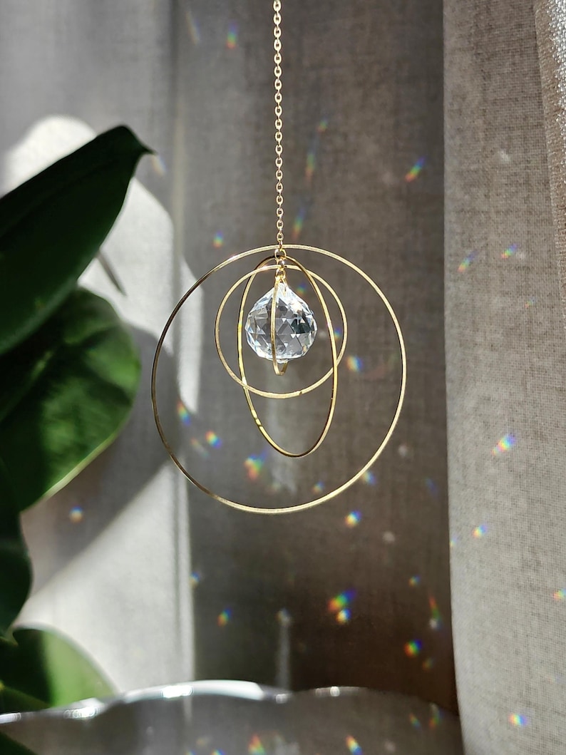 Un attrape-soleil suspendu composé d'une sphère en cristal transparent à facettes placée au centre de quatre anneaux métalliques dorés concentriques. L'ornement est fixé à une fine chaîne dorée, captant la lumière pour créer des reflets arc-en-ciel.