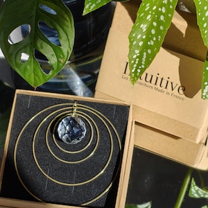 May include: A suncatcher featuring a faceted clear crystal sphere suspended within three concentric gold-toned metal rings. The item is presented in a square brown cardboard box with black foam padding, labelled with the text Intuitive Les Suncatchers Made in France.
