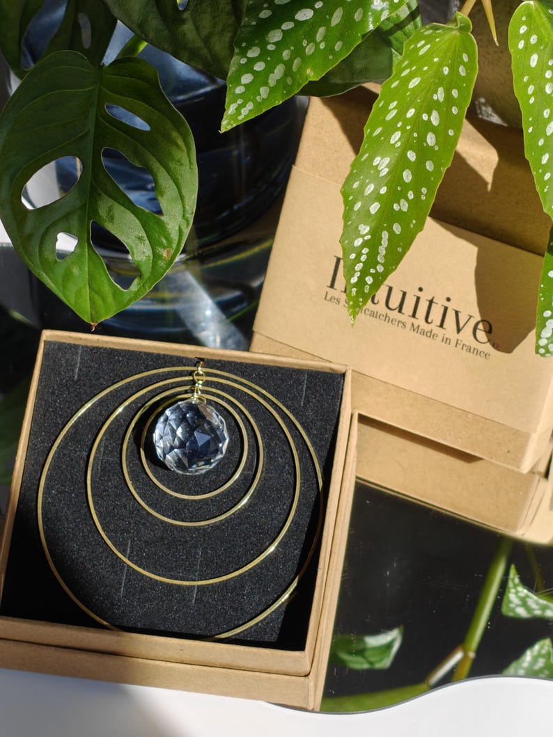 May include: A suncatcher featuring a faceted clear crystal sphere suspended within three concentric gold-toned metal rings. The item is presented in a square brown cardboard box with black foam padding, labelled with the text Intuitive Les Suncatchers Made in France.
