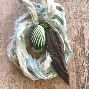 May include: A handmade necklace featuring a green and white striped ceramic pendant, a dark brown wooden element, and a light blue fabric tie. The necklace is displayed on a wooden surface.