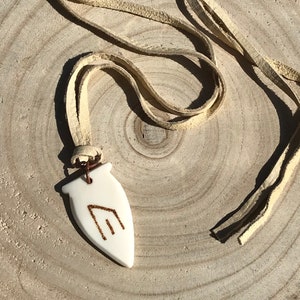 May include: A white ceramic pendant with a brown symbol on it, hanging from a light brown leather cord.