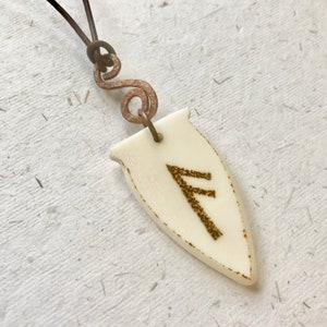 May include: A white ceramic pendant shaped like a shield with a brown rune symbol, "Fehu", etched on it. The pendant is attached to a brown leather cord with a copper wire loop.