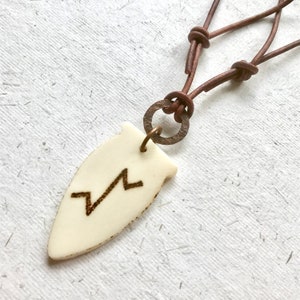 May include: A white bone pendant with a lightning bolt symbol carved into it. The pendant is attached to a brown leather cord with a brass ring.