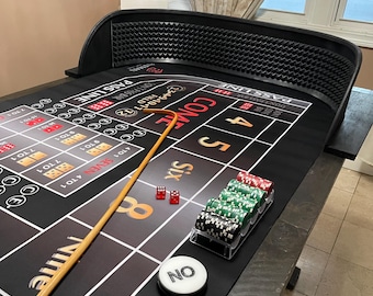 Puede incluir: Una mesa de craps con una superficie de fieltro negro, con las palabras "PASS LINE" y "COME". Dados rojos, fichas de casino y un palo de madera están sobre la mesa. También es visible un marcador blanco "NO".