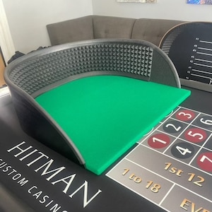 May include: A custom casino table with a green felt playing surface and a black frame. The table features the text "HITMAN CUSTOM CASINO" in white. The table also has a roulette wheel and betting layout.