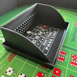 May include: A black craps dice tray with a felt interior, featuring white text and a red die. The tray sits on a green craps table with dice and betting areas. The text on the tray includes "COME", "PASS LINE", and numbers.