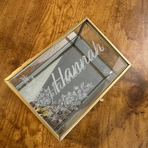 May include: A rectangular glass jewelry box with a gold metal frame and a hinged lid. The lid features an etched floral design and the name Hannah in script lettering. The interior base is lined with a grey fabric, and a small gold chain is visible.