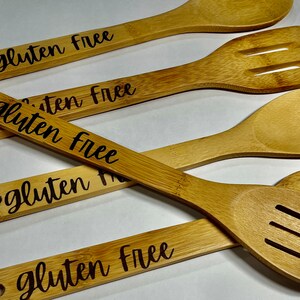 Peut inclure: Ensemble de quatre cuill&egrave;res en bois avec les mots "sans gluten" grav&eacute;s sur chacune. Les cuill&egrave;res sont en bambou naturel et ont une couleur brun clair.
