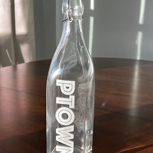 May include: A clear glass bottle with a white lid and a metal swing top. The bottle has the word "PTOWN" etched on the side.