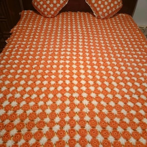 May include: Handmade orange and white crochet blanket with matching square cushions. The blanket and cushions feature a repeating floral pattern, creating a cosy and decorative bedding set.