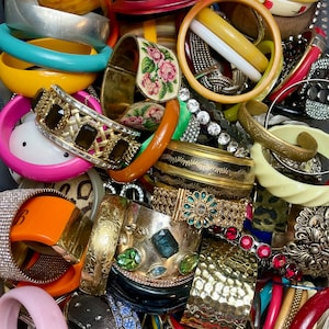 May include: A large collection of colorful and decorative bangles, bracelets, and cuffs. Many of the bangles are made of plastic, while others are made of metal and feature intricate designs. Some of the bangles are adorned with rhinestones and other embellishments.